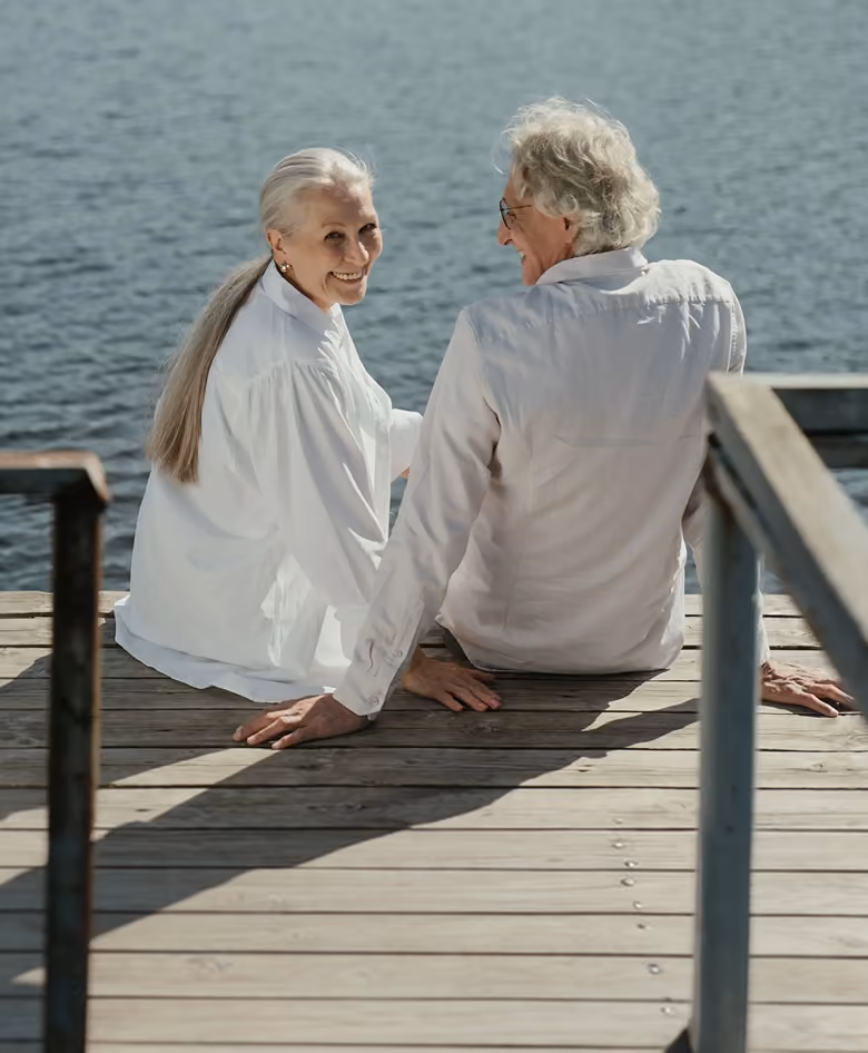 Elderly couple standing close together by a lake with a forest and clear blue sky in the background, holding hands and smiling at each other.