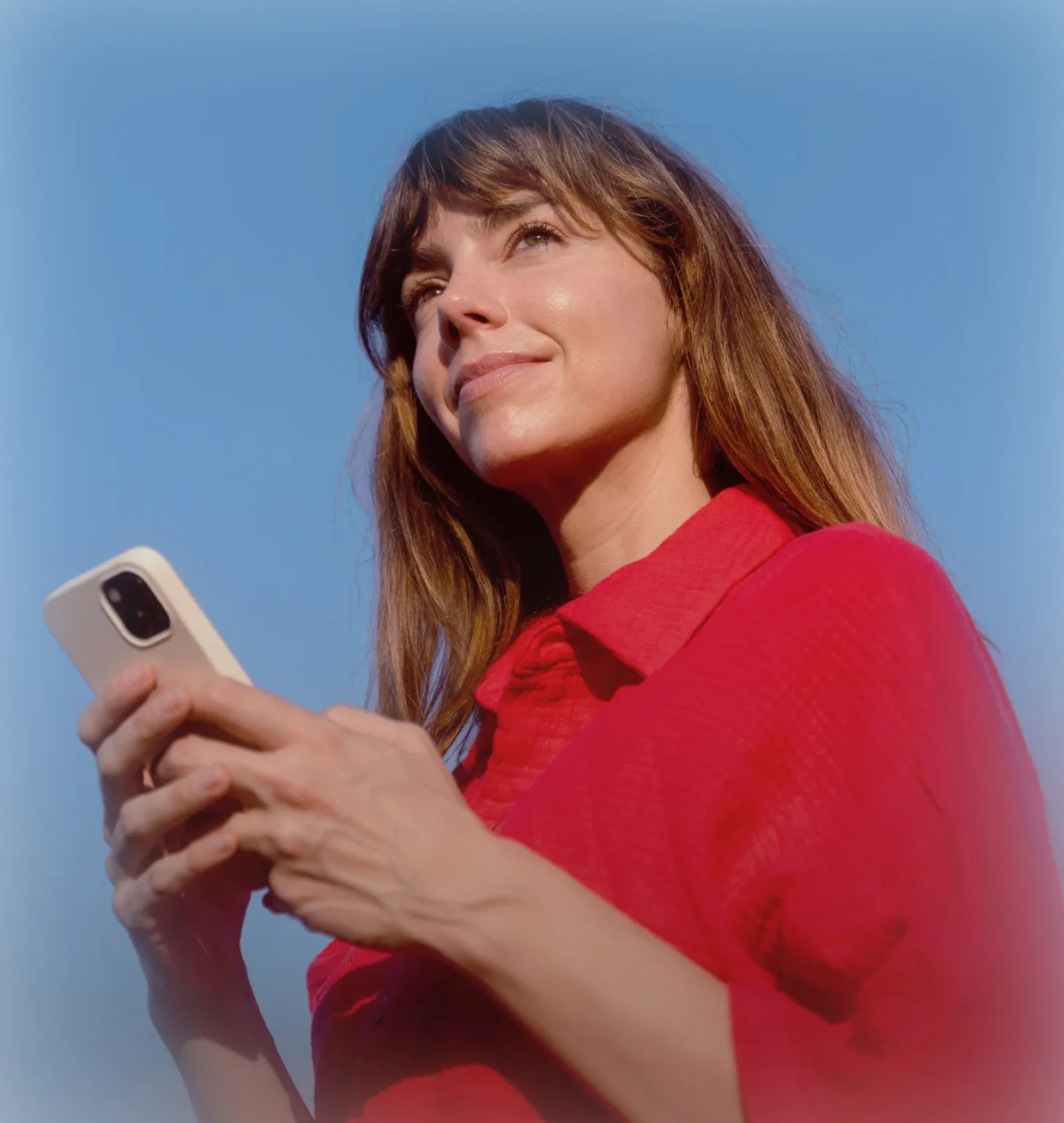 Smiling woman in a red shirt holding a phone and looking up against a blue sky