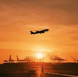 Plane taking off at sunset over a port with container cranes