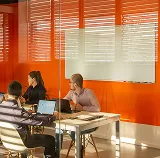 Three colleagues meeting at a table beside a whiteboard in an orange-walled office