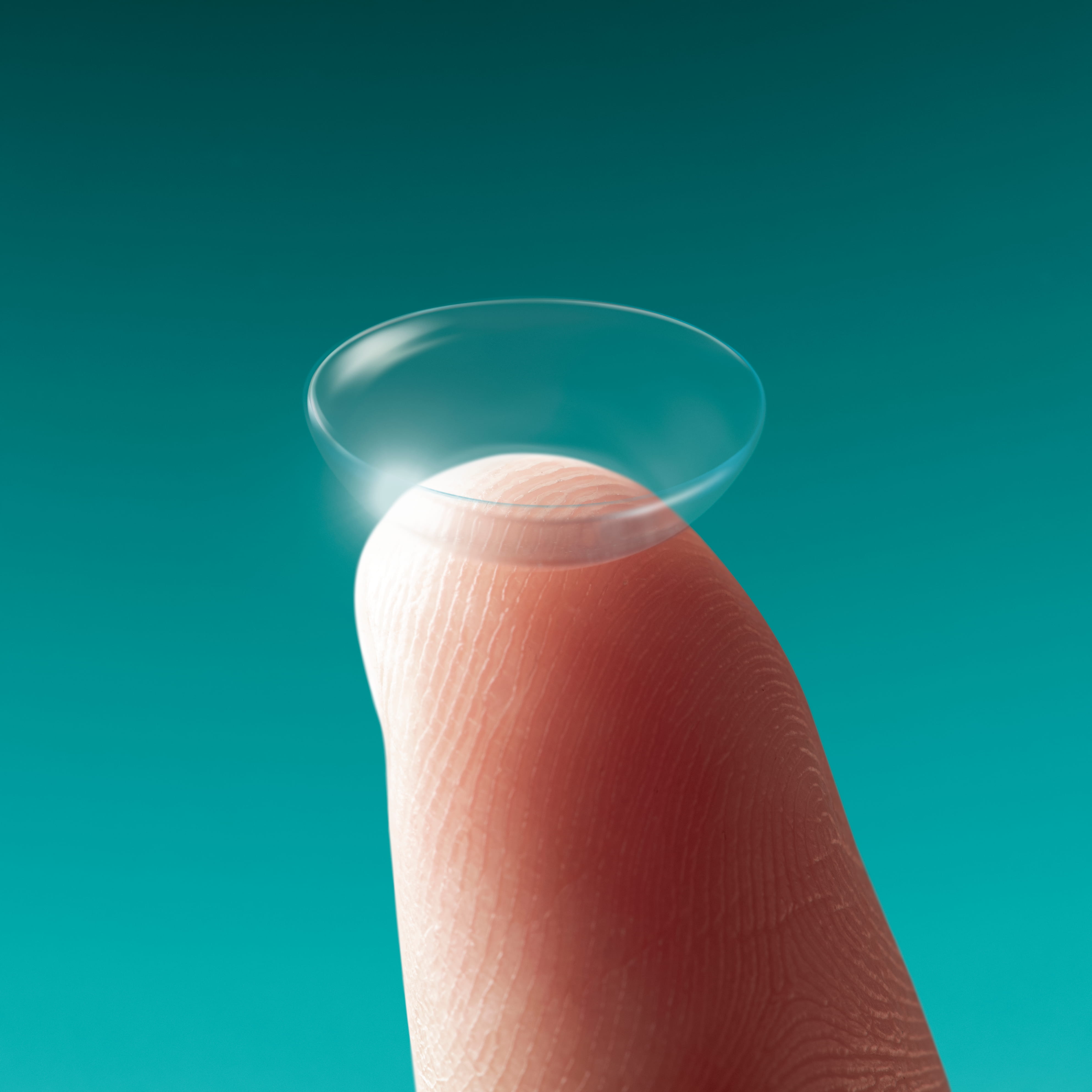 A transparent contact lens balanced on the fingertip of a person against a teal background.