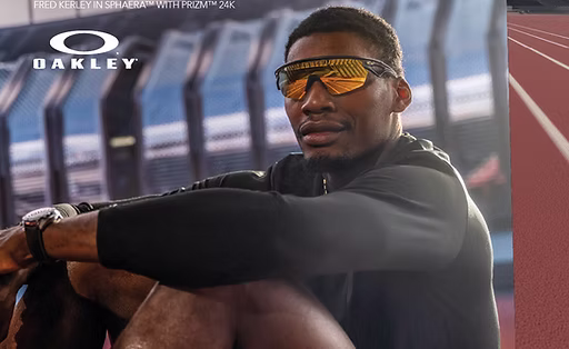 An athlete wearing reflective Oakley sunglasses and a black jacket sits relaxed inside a sports stadium near a running track.