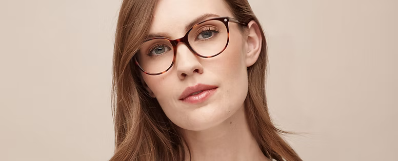 Close-up portrait of a woman with light skin, light brown hair, wearing tortoiseshell glasses, against a beige background.