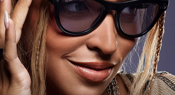 Close-up of a smiling woman wearing black Ray-Ban sunglasses with braids and styled hair, slightly touching the frame.