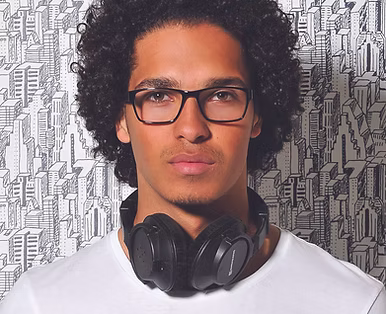 Young man with curly hair wearing glasses and a white shirt, with black headphones around his neck, standing in front of a black and white cityscape illustration background.