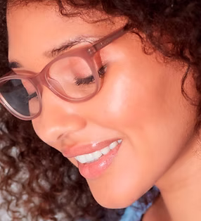 Close-up of a smiling woman with curly hair wearing pink translucent eyeglasses, looking downward.