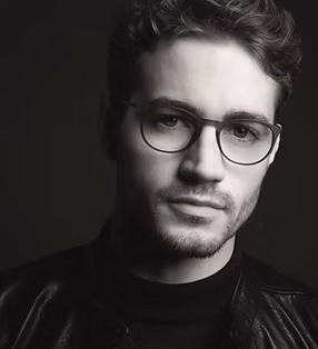 Black-and-white portrait of a young man with short wavy hair, wearing round glasses, a dark turtleneck, and a leather jacket, looking slightly to the side against a dark background.