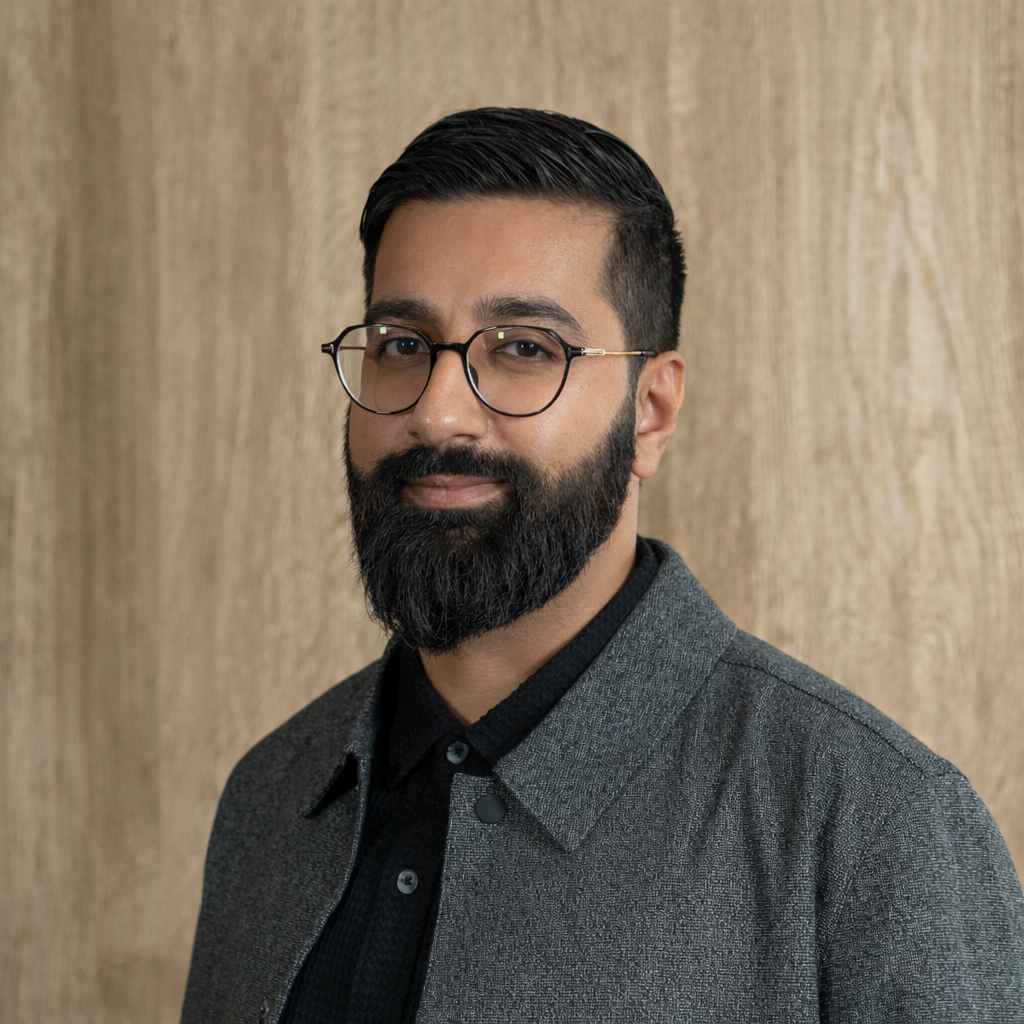 Man with dark hair and full beard, wearing round glasses, a black shirt, and a gray jacket, posing against a light wooden background.