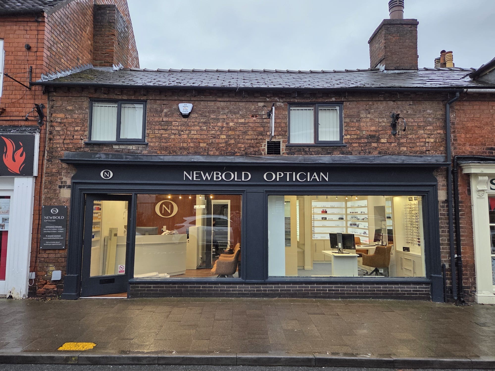 Exterior view of Newbold Optician shop with dark framing, two front windows showing interior seating and display of eyeglasses, and two upper-floor windows on a brick building.