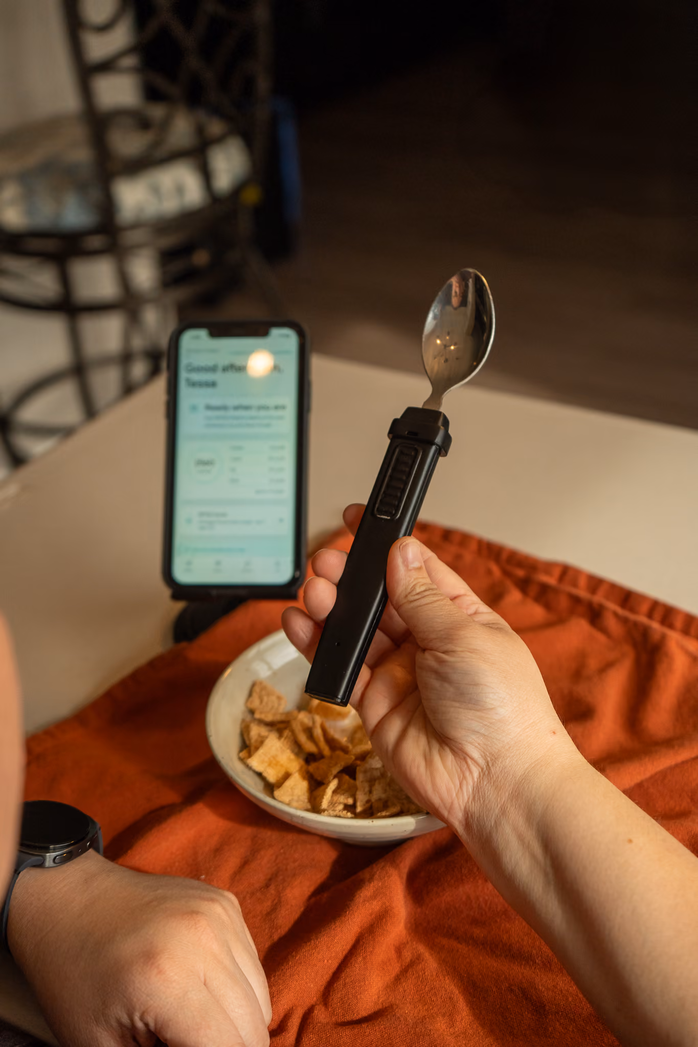 Person at a dining table holding the BiteQ utensil beside a bowl of food and a phone, ready to pair it