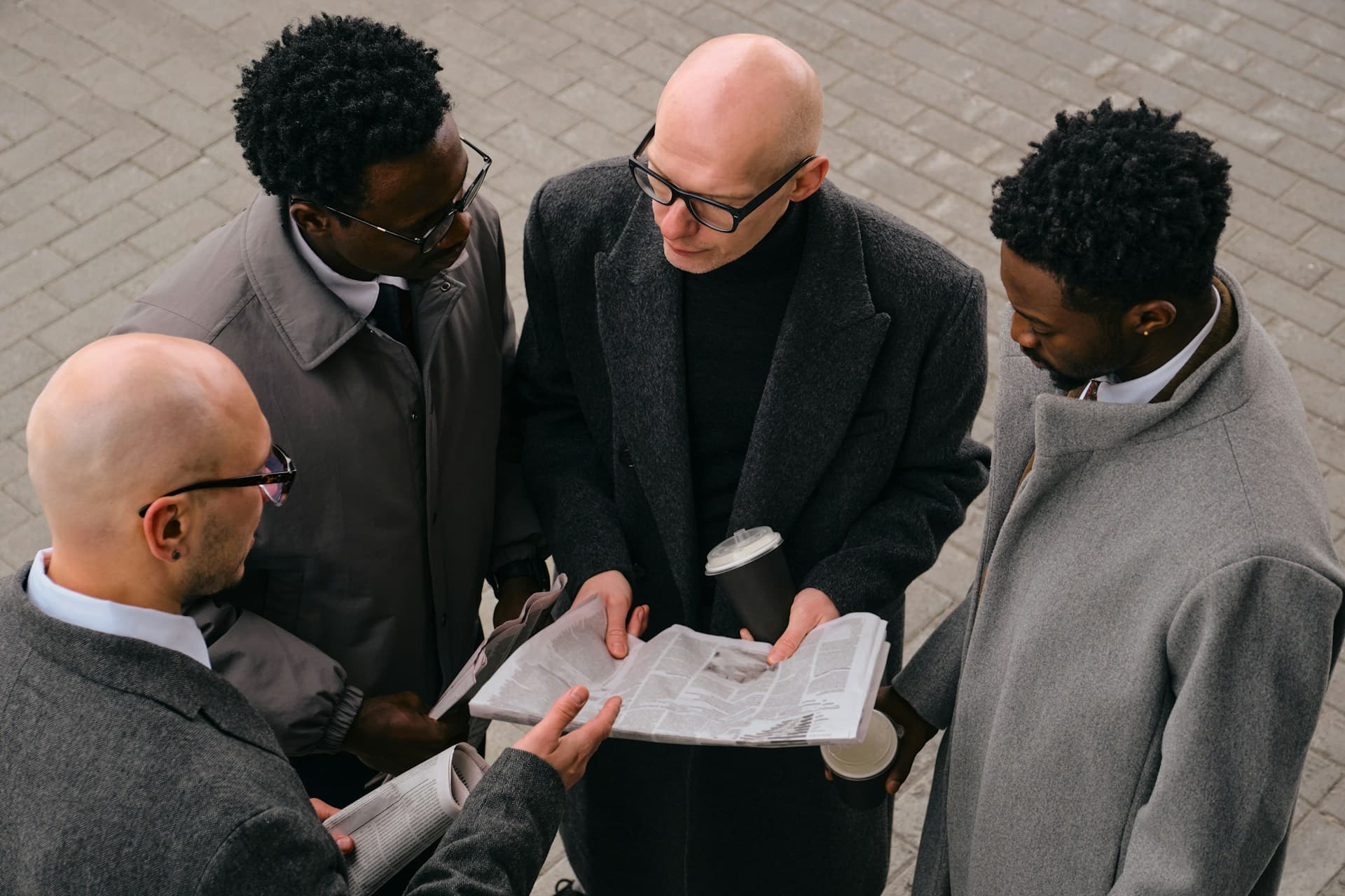 Four men in coats standing outdoors on a paved surface, holding coffee cups and newspapers, engaged in conversation while looking at the newspaper.