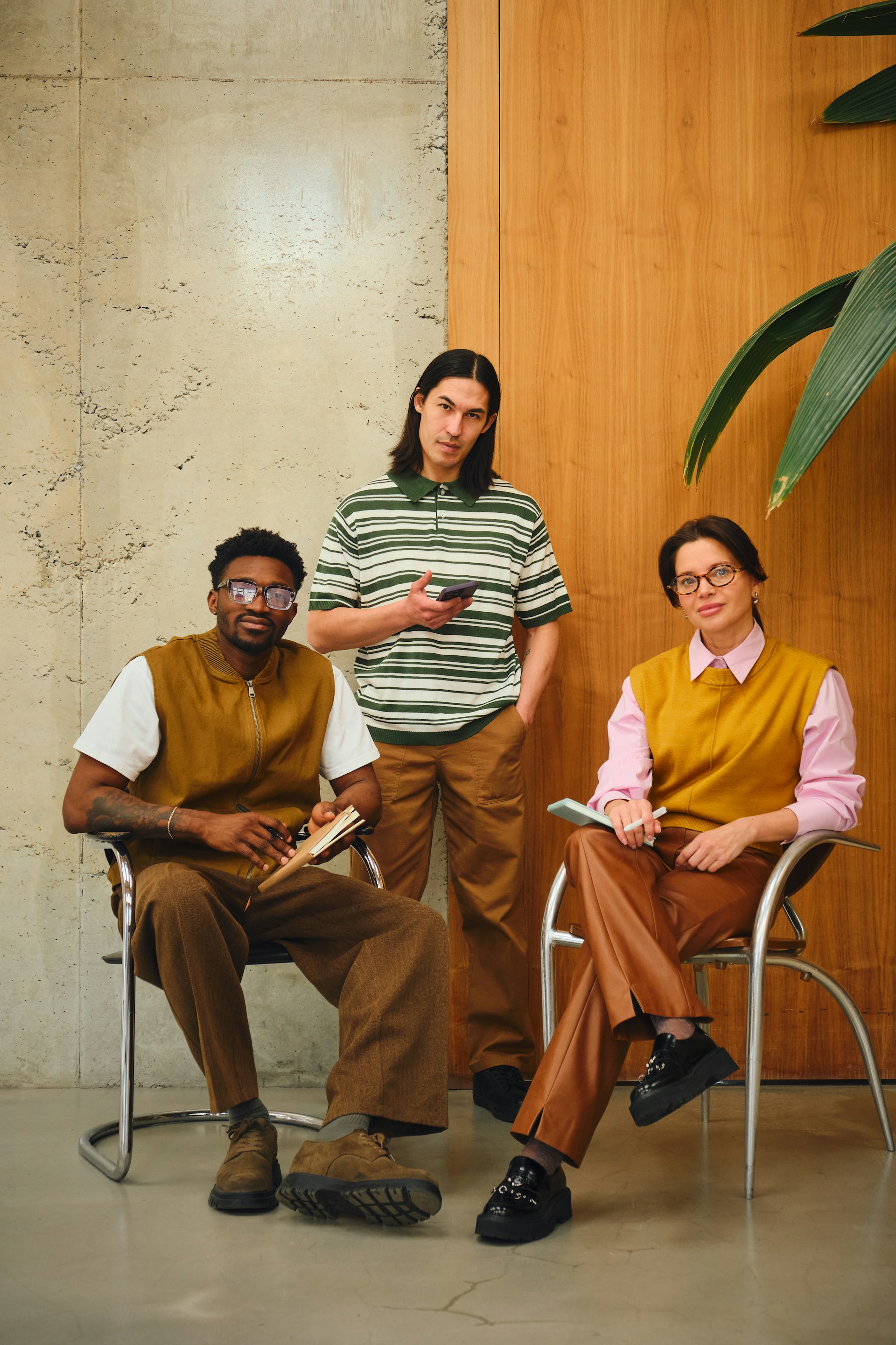 Three people in a modern indoor setting with concrete and wood-paneled walls; one man with glasses and a brown vest sits on a chair holding a notebook, another man in a green and white striped shirt stands holding a phone, and a woman with glasses, wearing a yellow vest and brown pants, sits on a chair holding a pen and notebook.