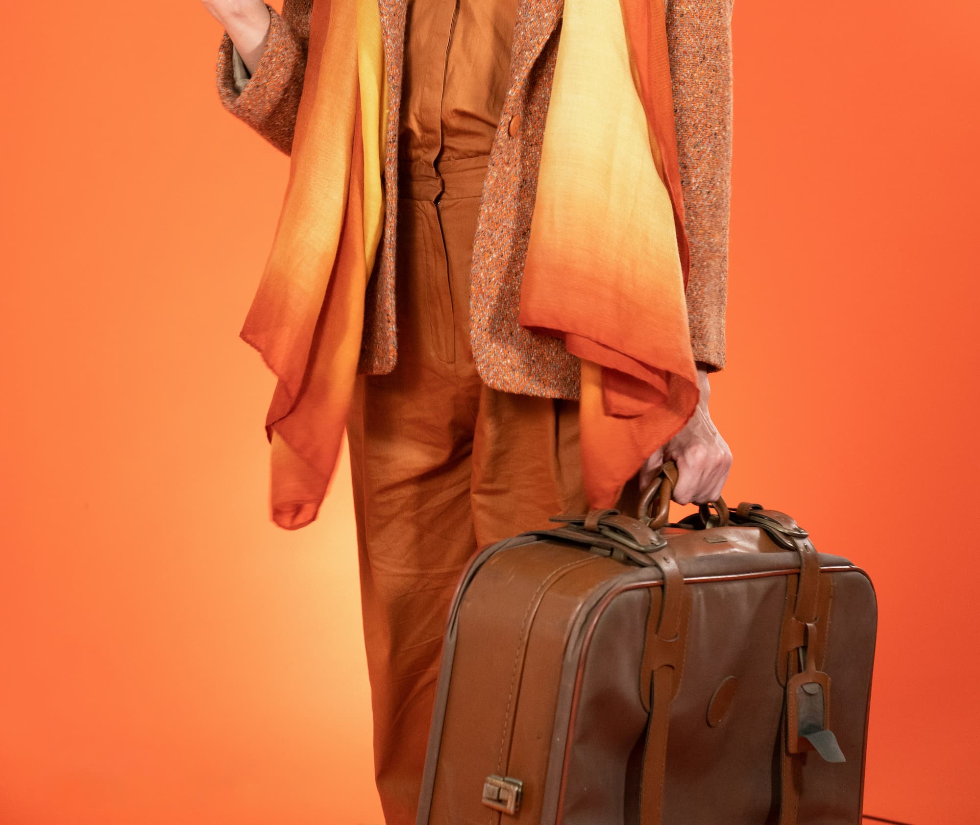 Person wearing earth-toned clothes and an orange-yellow gradient scarf holding a brown leather suitcase against an orange background.