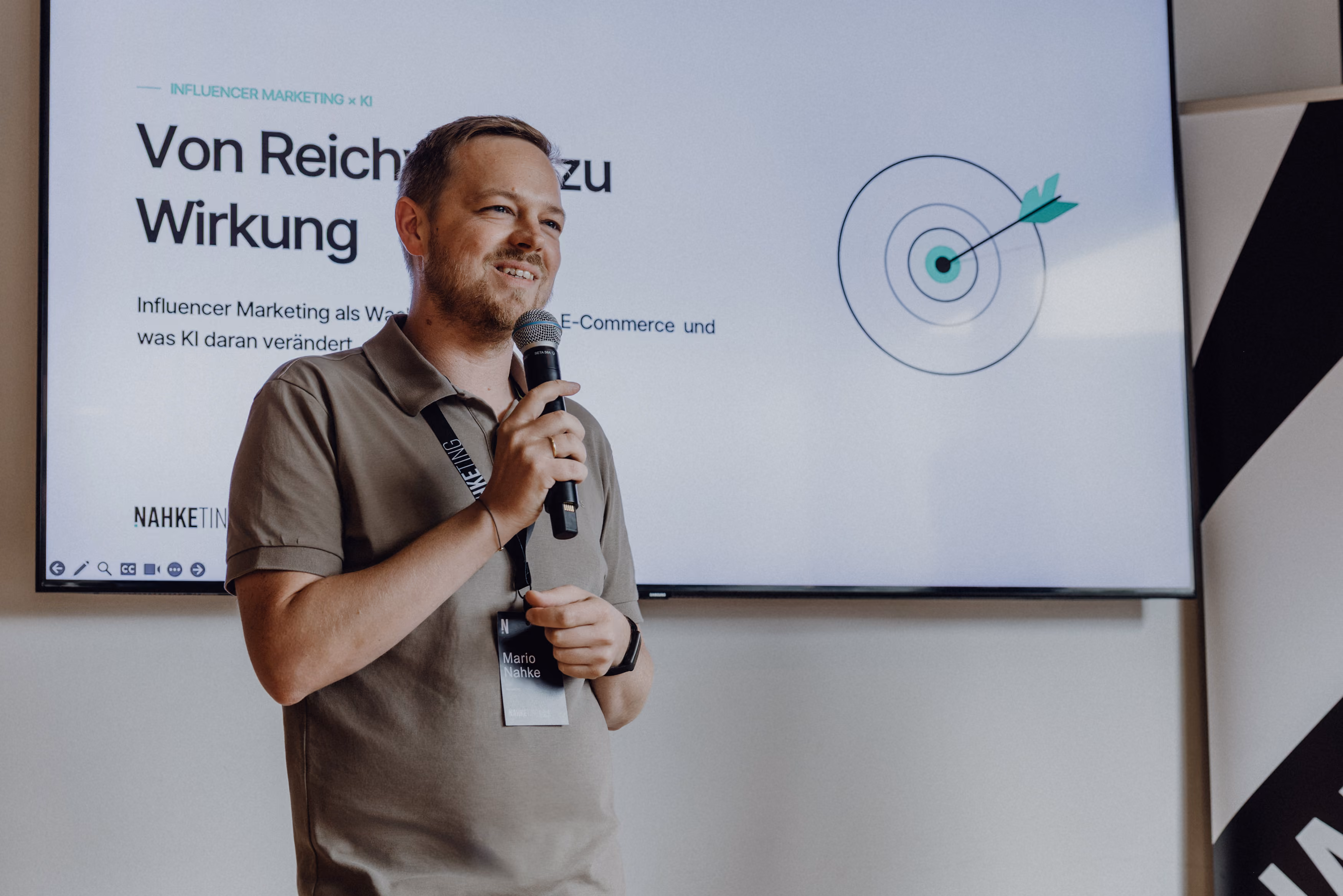 Ein Mann, der ein Mikrofon in der Hand hält und lächelt, während er vor einer Leinwand einen Vortrag hält, auf der eine Zielscheibe mit einem Pfeil im Zentrum und ein Text über Influencer-Marketing auf Deutsch zu sehen ist.