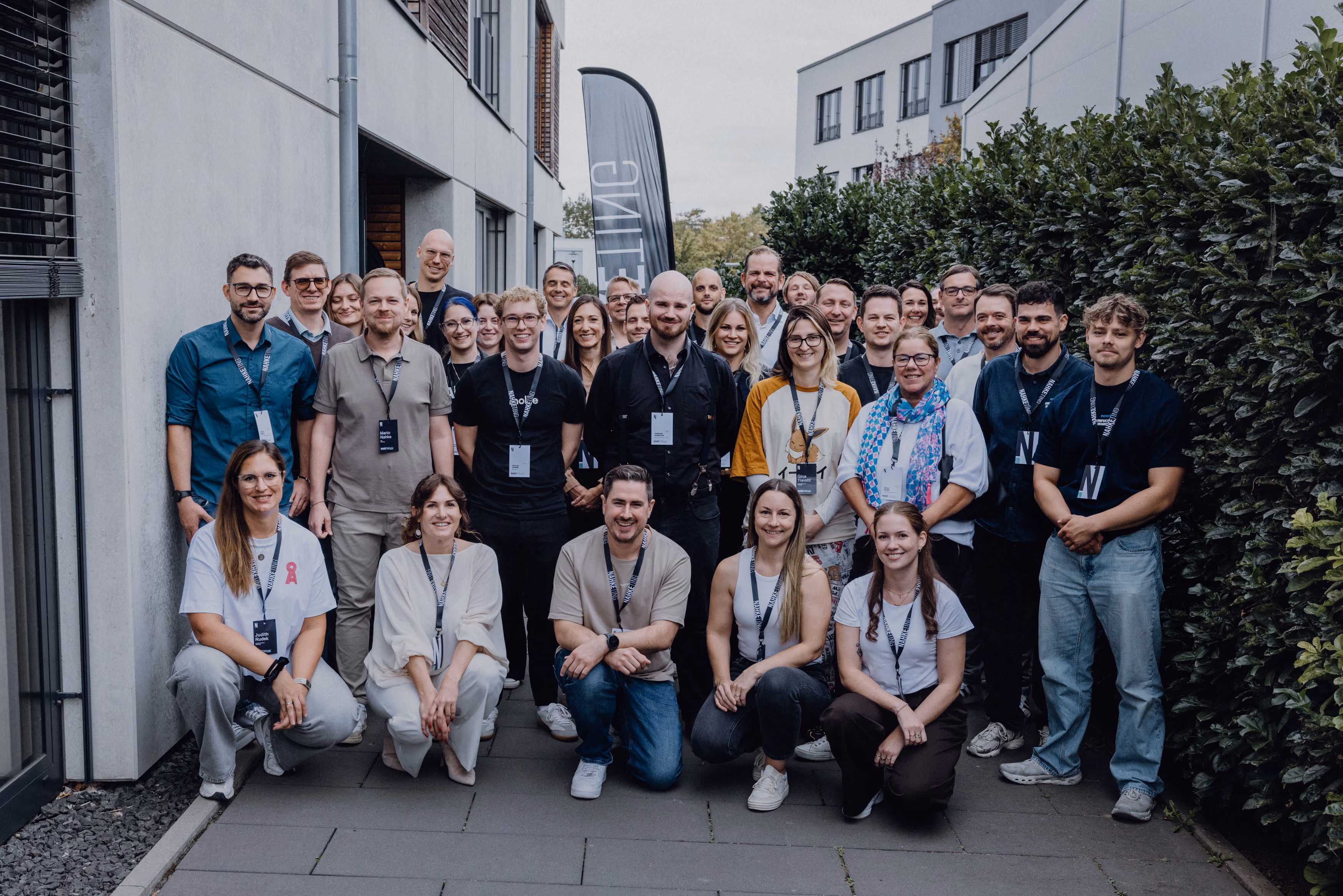 Gruppenfoto von etwa 40 Personen, die im Freien zwischen Gebäuden und Büschen stehen und knien; viele tragen Umhängebänder mit Namensschildern und lächeln in die Kamera.