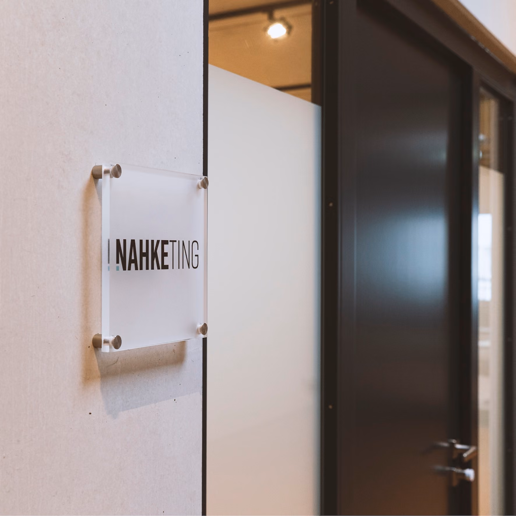 Transparentes, quadratisches Türschild mit der Aufschrift „NAHKETING“, angebracht an einer hellen Wand neben einer teilweise sichtbaren schwarzen Tür.