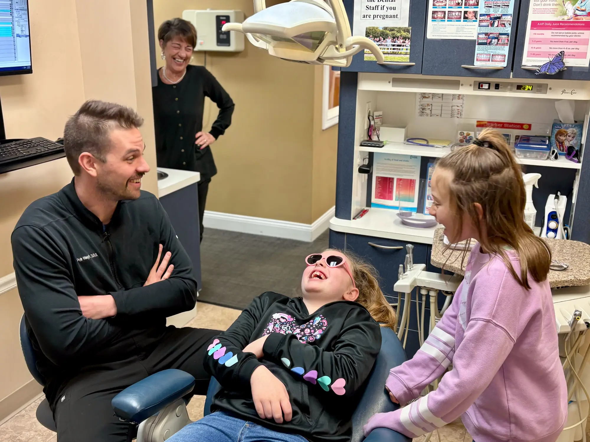 Dentist smiling and talking with two young girls in a dental office, one girl reclining in the dental chair wearing sunglasses and laughing.