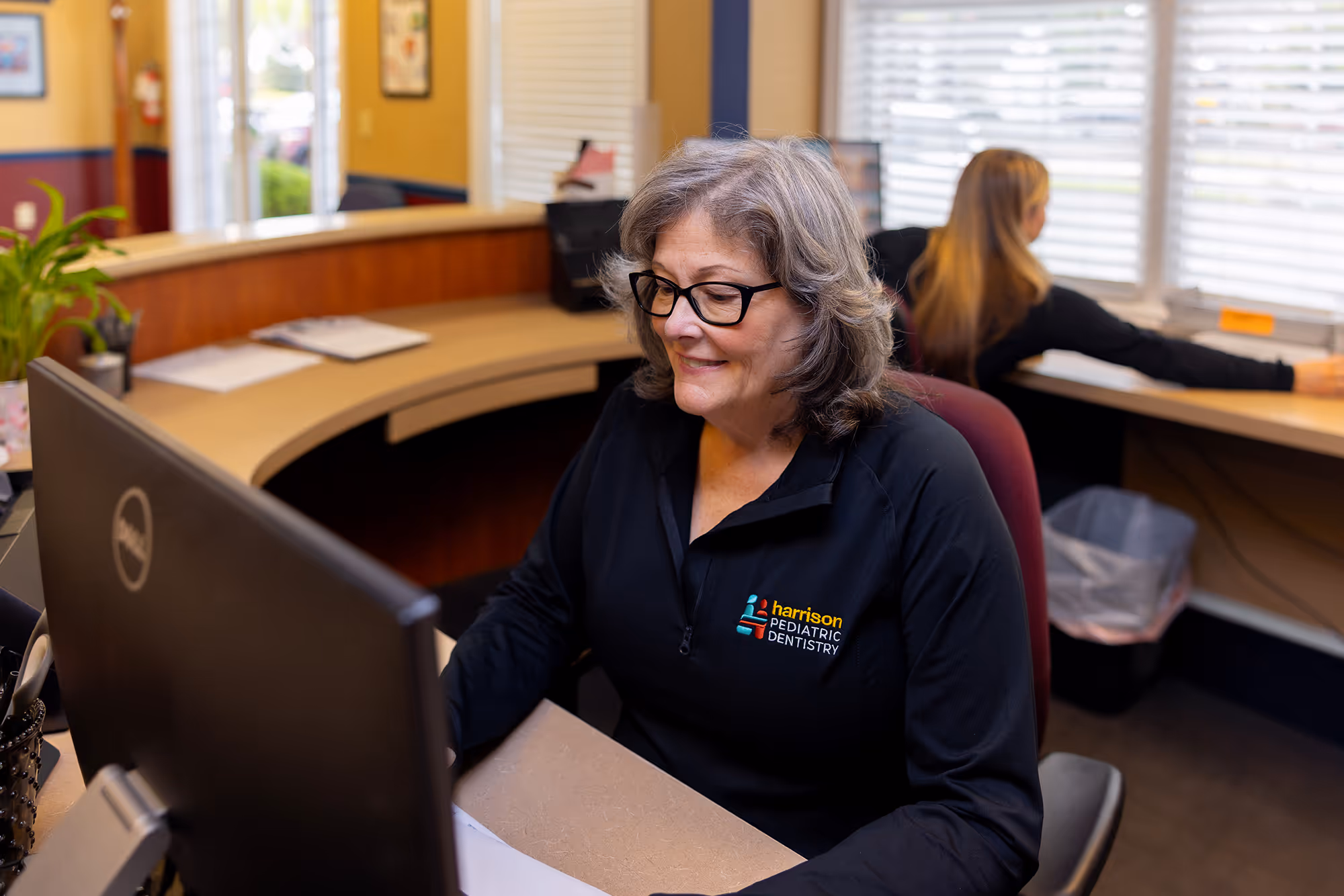 Smiling woman with glasses working at a computer in a dental office wearing a black jacket with Harrison Pediatric Dentistry logo.