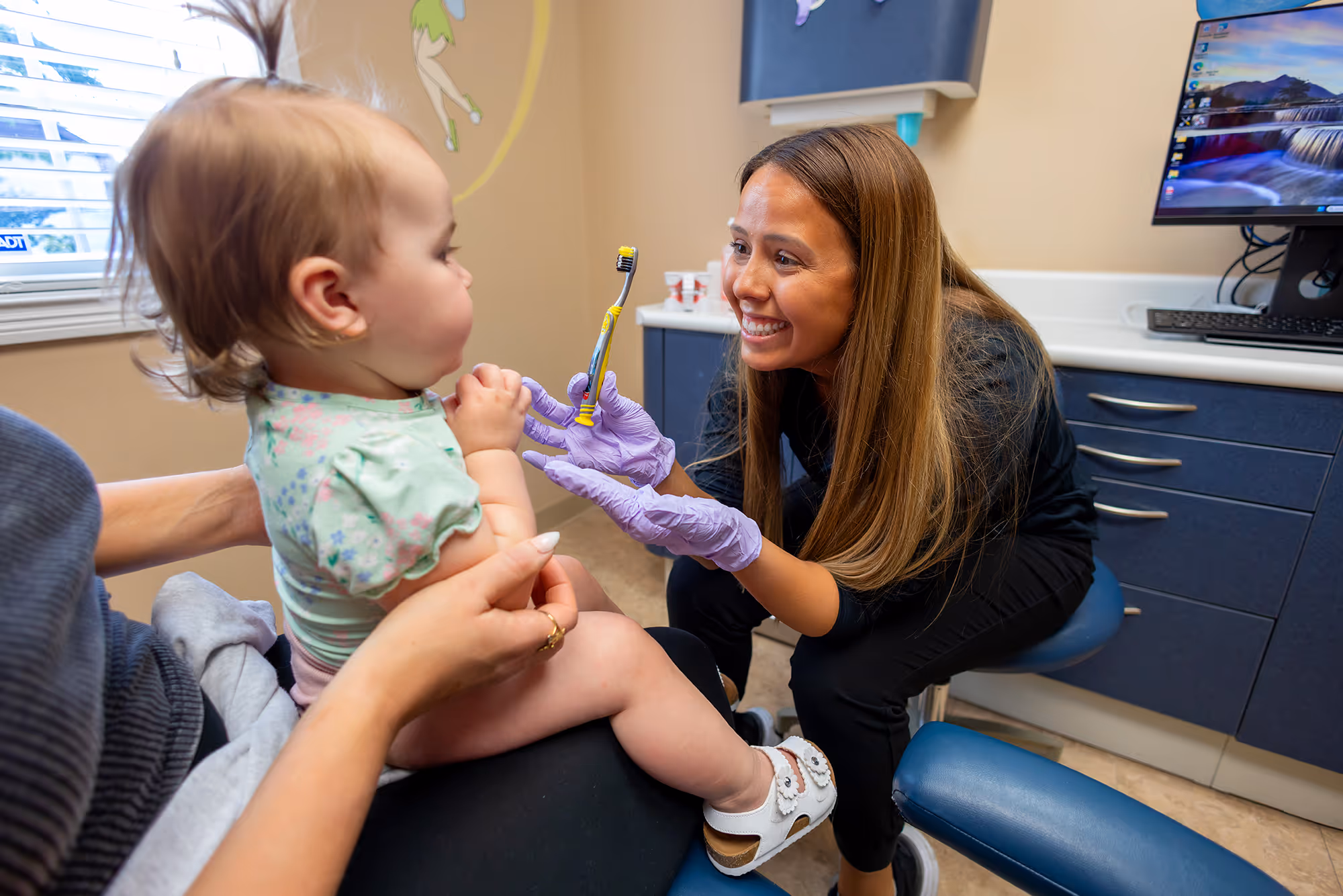 Smiling dental hygienist wearing purple gloves showing a toddler a toothbrush in a dental office.