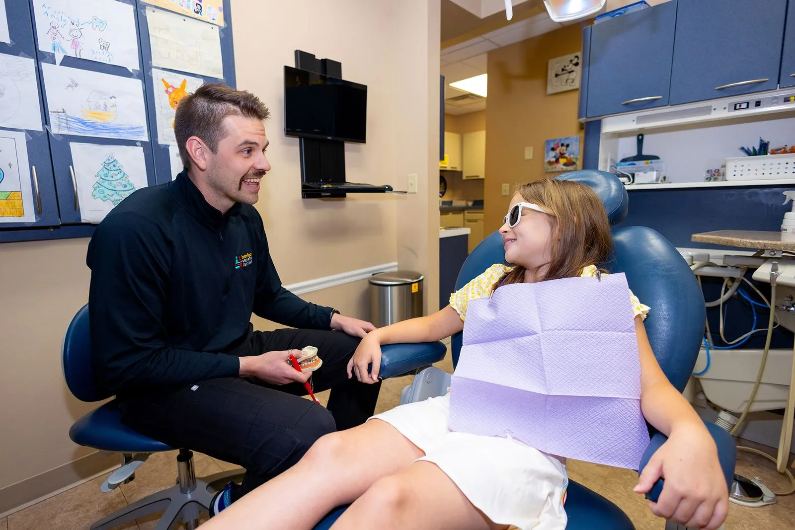 Dentist demonstrating teeth brushing on a model to a young girl wearing sunglasses sitting in a dental chair.