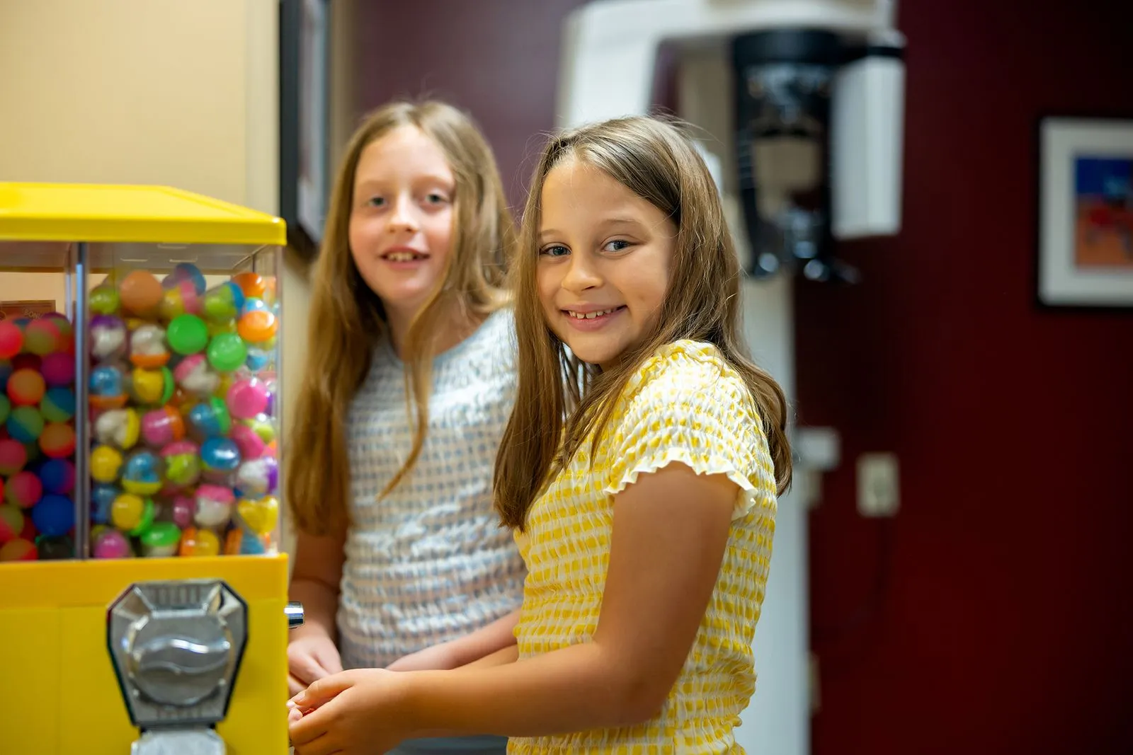 Two young girls smiling and standing next to a yellow gumball machine filled with colorful gumballs inside the best pediatric clinic in Ohio.