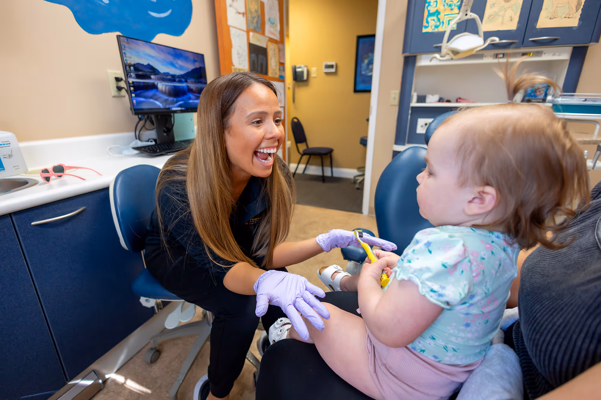 Smiling female dental hygienist wearing purple gloves engaging with a toddler holding a yellow toothbrush in a dental office.