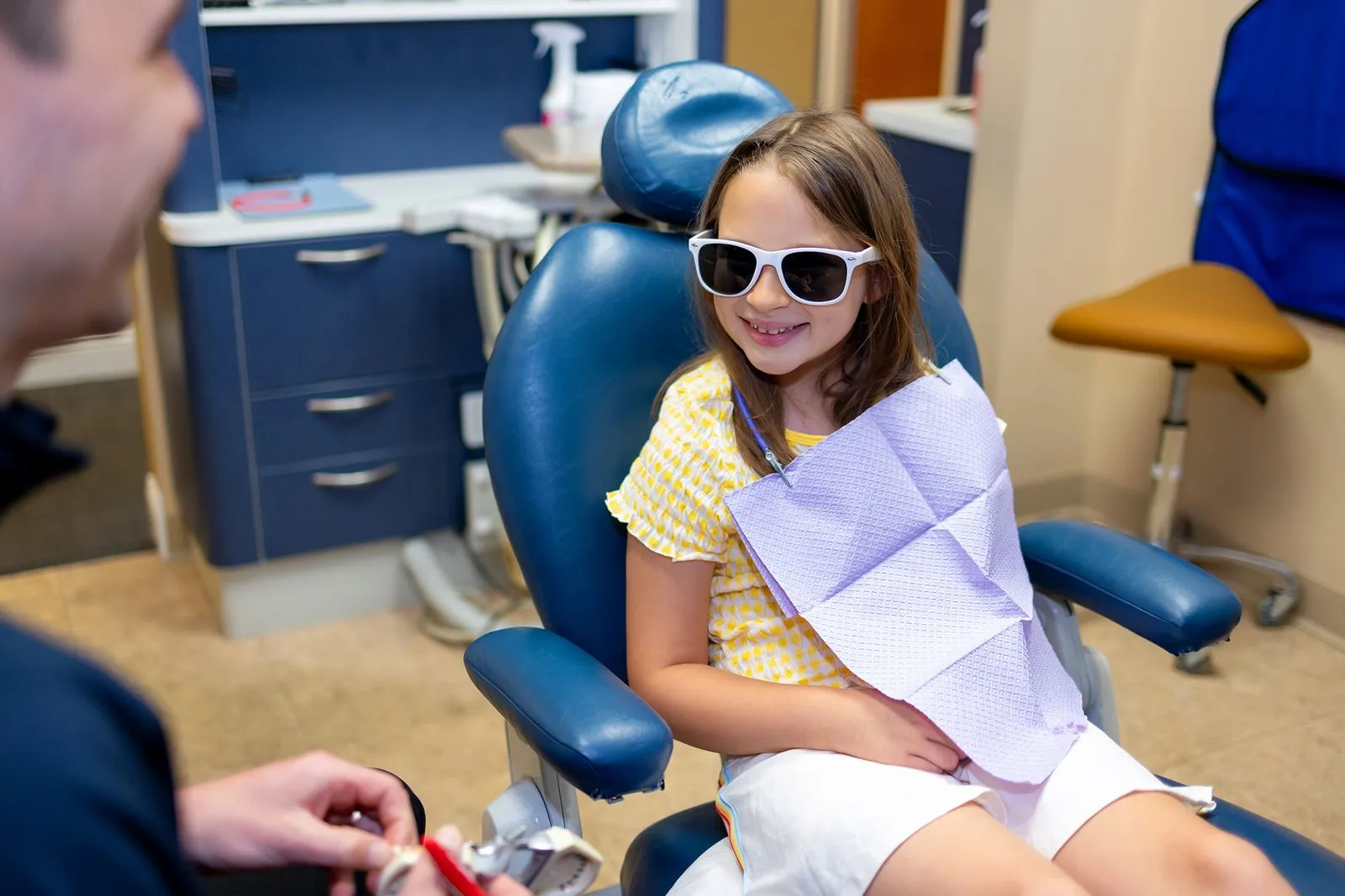 Smiling young girl wearing sunglasses sitting in a dental chair with a dental bib, facing a person holding a dental instrument.