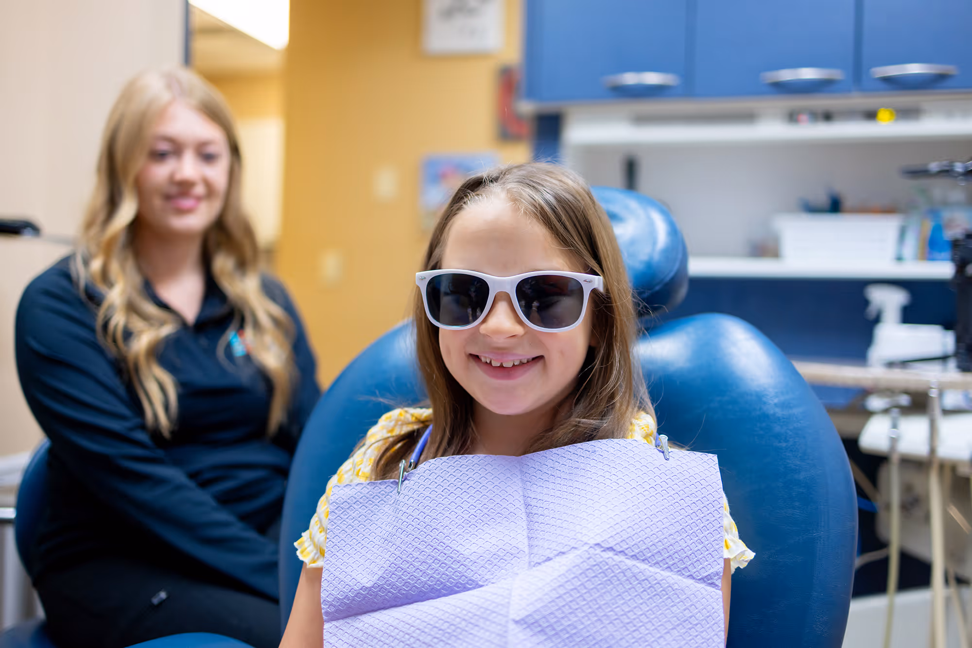 Smiling girl wearing sunglasses sitting in a dental chair with a dental bib, dental professional in background.