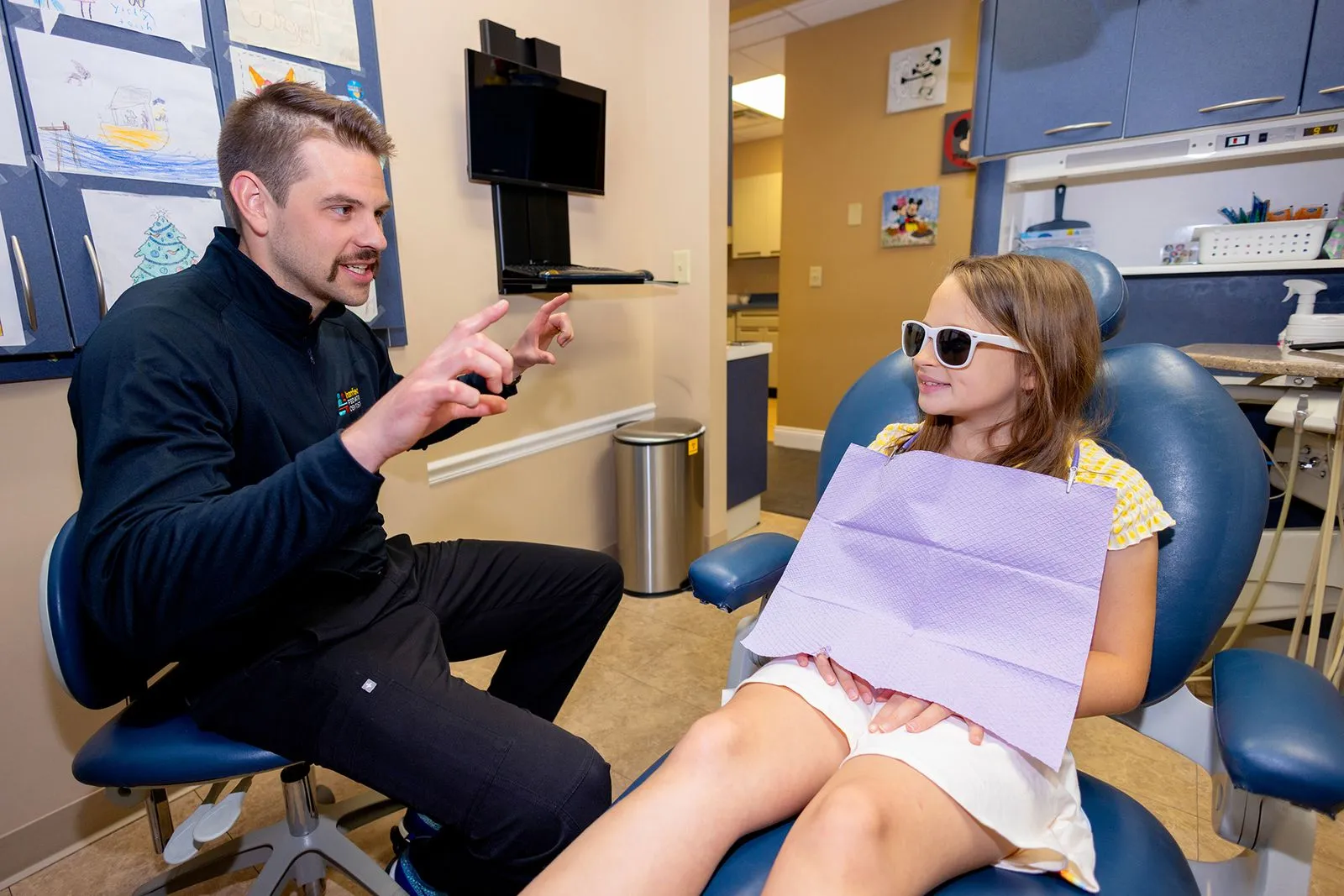 Dentist playfully gestures while talking to a smiling young girl wearing sunglasses in a dental chair.