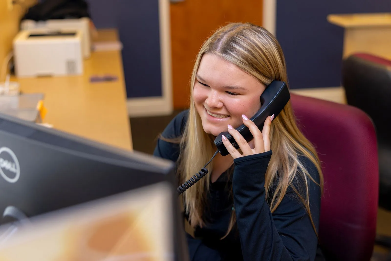 Smiling woman with long blonde hair speaking on a desk phone while looking at a computer monitor in an office setting.