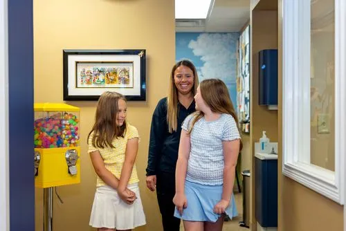 Two young girls smiling and walking with a woman in a black jacket inside an office hallway with a colorful gumball machine on the left.