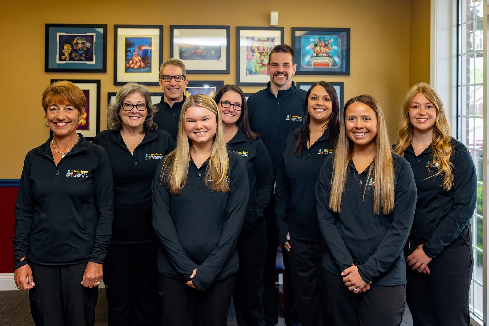 Group photo of nine smiling dental team members wearing black uniforms with Harrison Pediatric Dentistry logo, standing indoors against a wall with framed artwork.