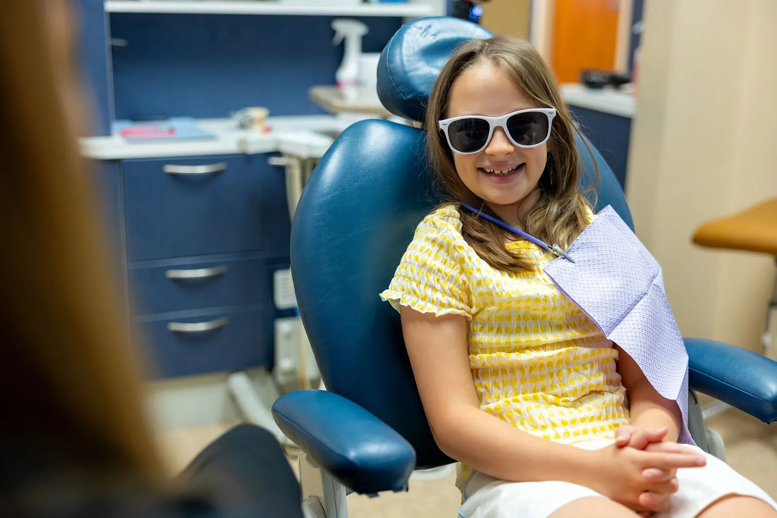 Smiling girl wearing sunglasses and yellow patterned shirt sitting in a dental chair with a dental bib.