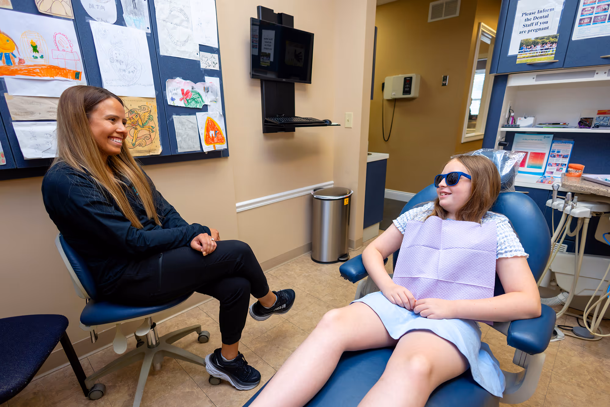 Young girl wearing sunglasses sitting in a dental chair smiling at a female dental professional.