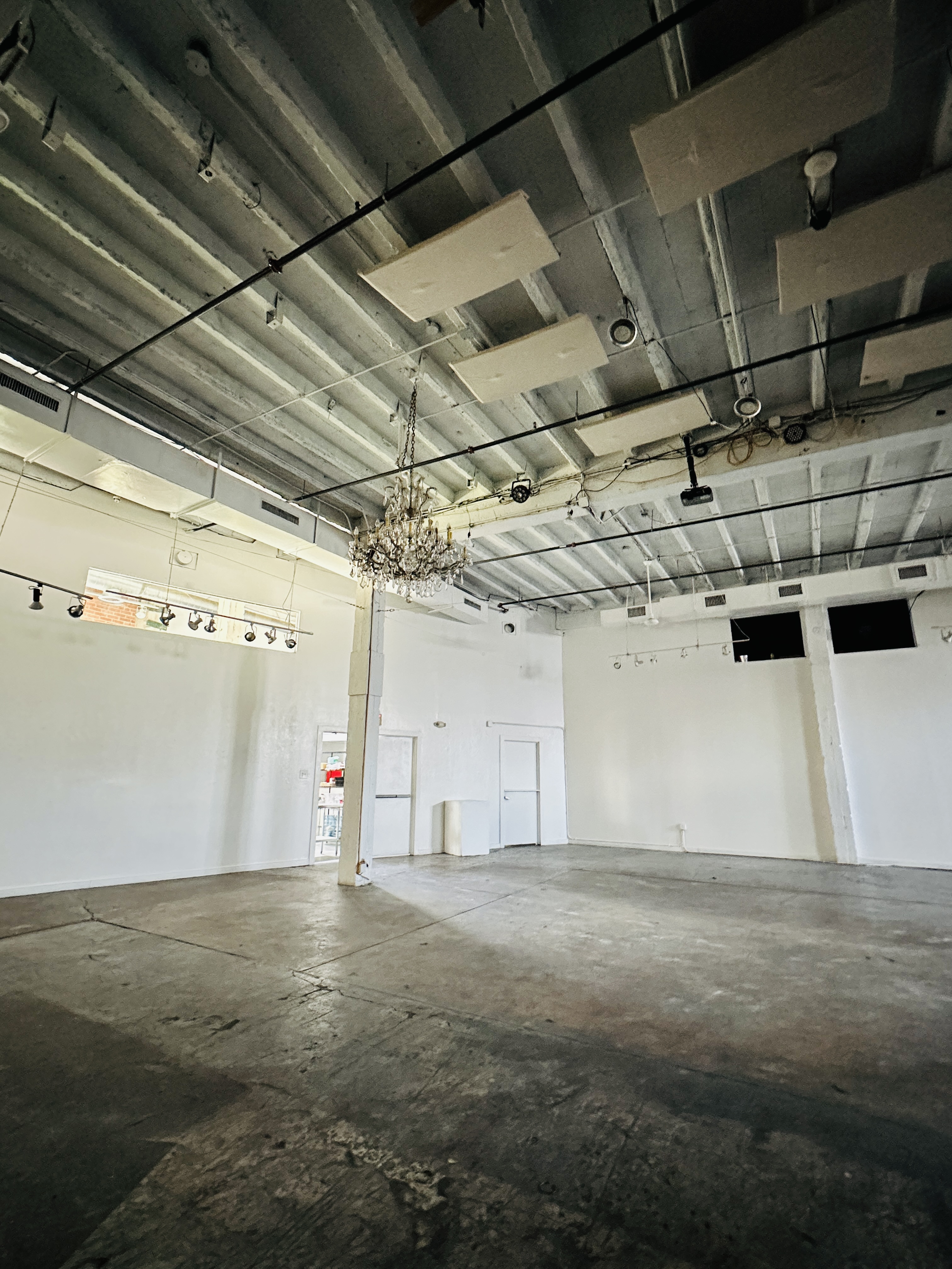 Empty industrial-style room with concrete floor, exposed ceiling beams, a hanging crystal chandelier, white walls, and a few doors and windows.