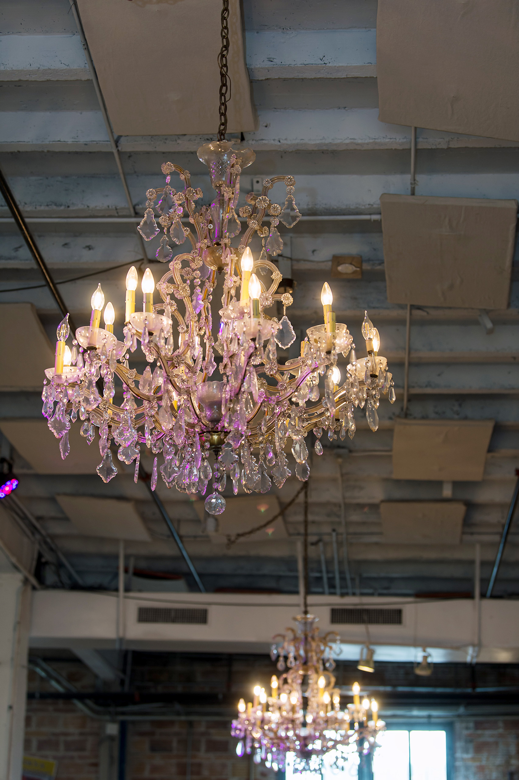 Two ornate crystal chandeliers hanging from a gray industrial ceiling with light bulbs lit, casting a soft glow.