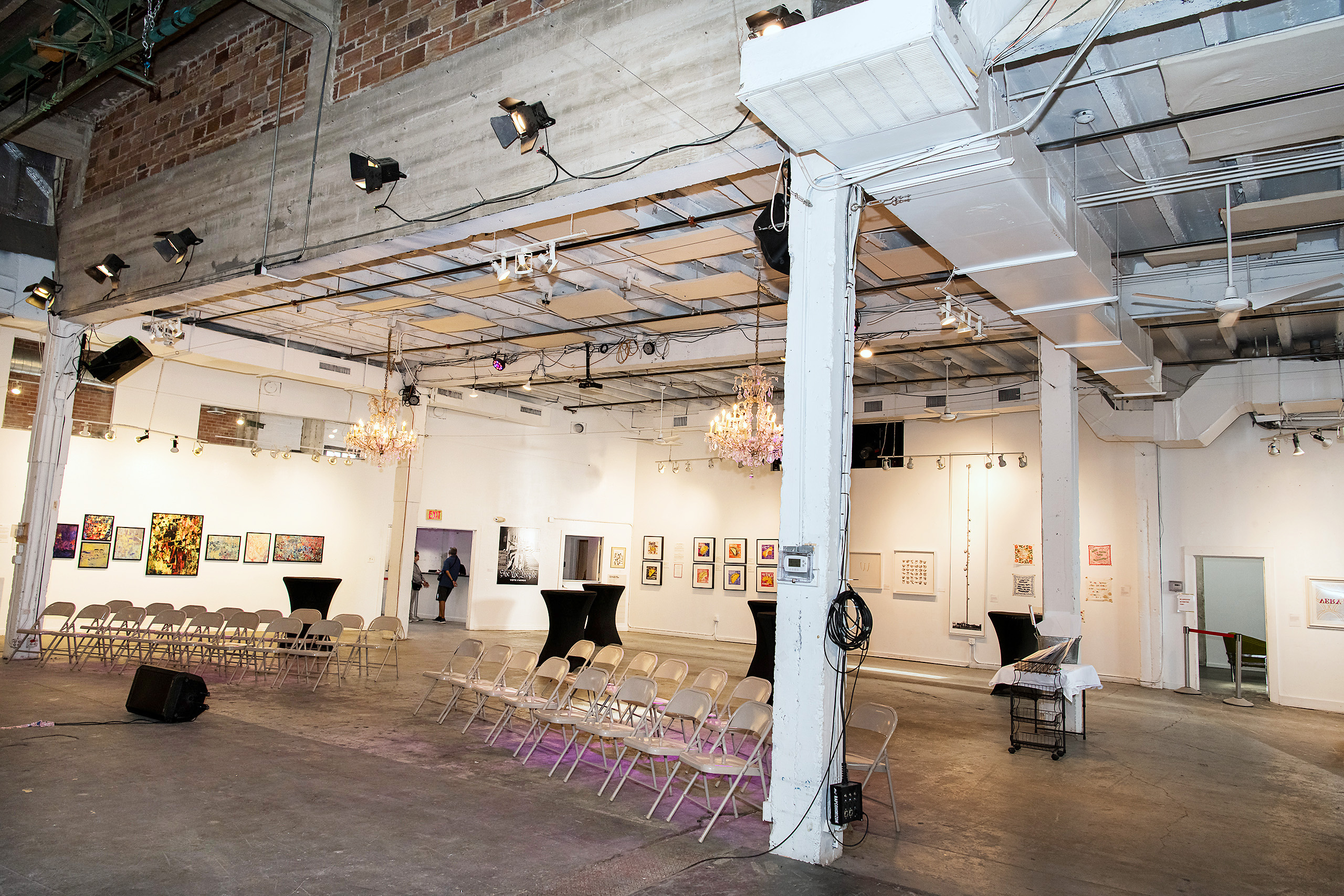 Interior of a contemporary art gallery with white walls displaying framed artworks, two rows of folding chairs arranged facing inward, a speaker on the floor, and ornate chandeliers hanging from the ceiling.
