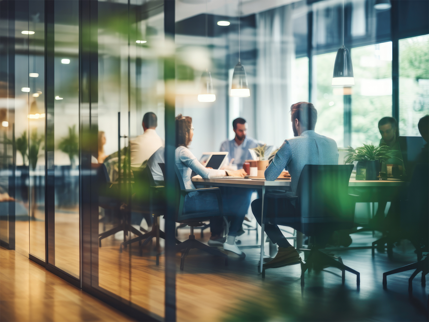 A group of people sitting around a conference table in a modern office with glass walls, working on laptops and discussing.