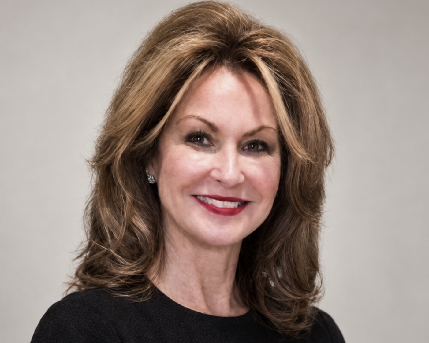 Smiling woman with shoulder-length light brown hair wearing a black top and small earrings against a plain background.