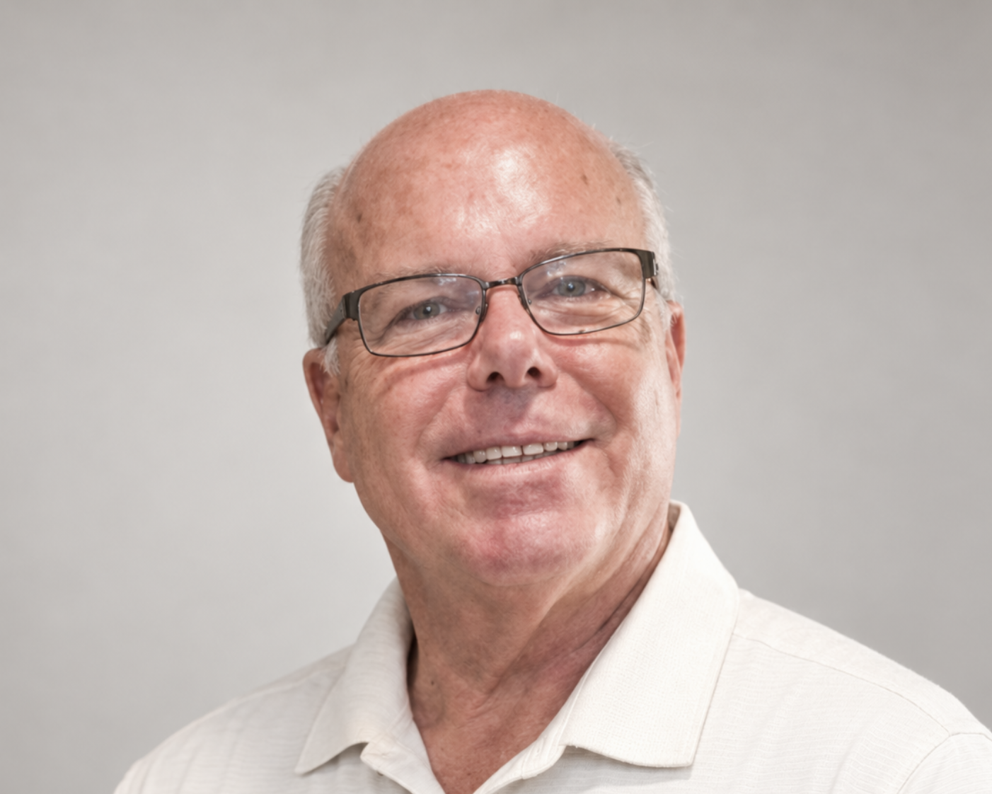 Smiling man with glasses, short white hair, and a light beige collared shirt against a plain gray background.