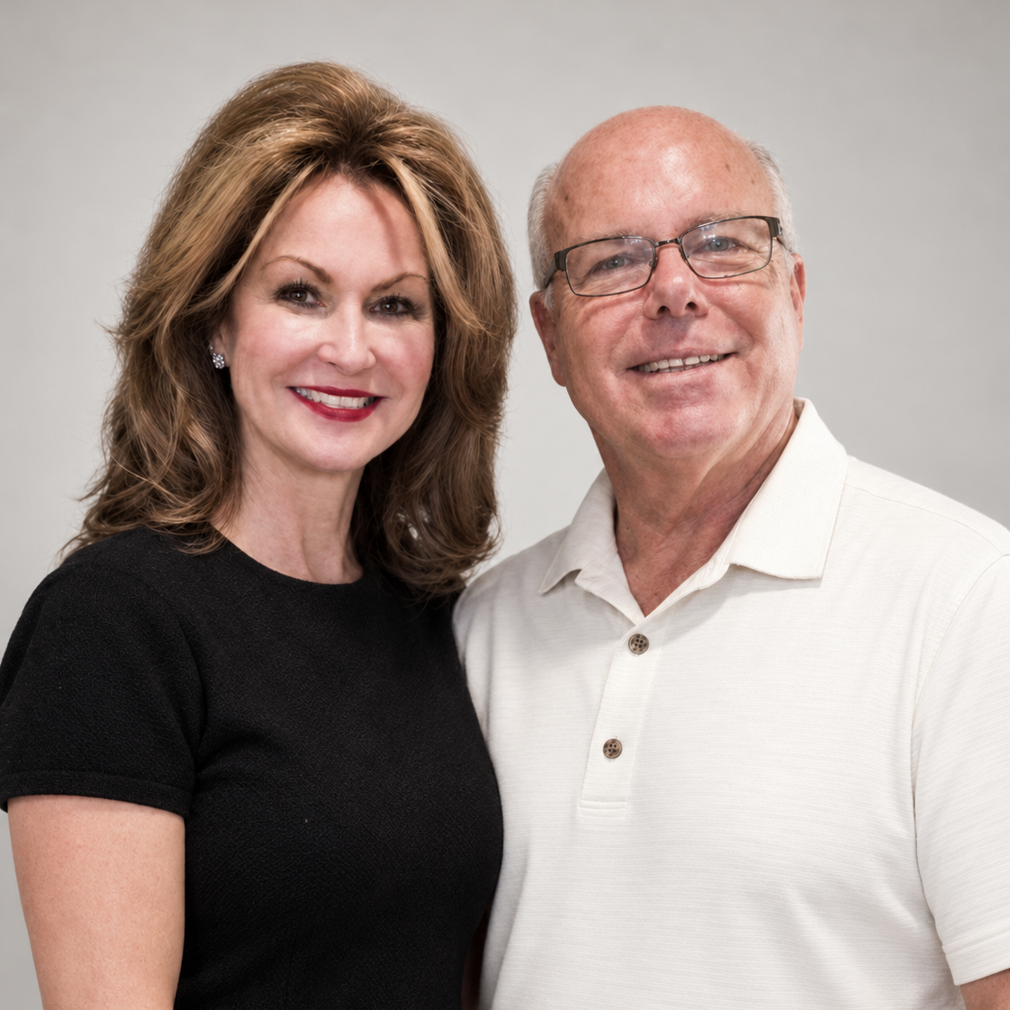 Smiling woman with styled brown hair wearing a black top stands next to a smiling man with glasses and a white polo shirt against a plain gray background.