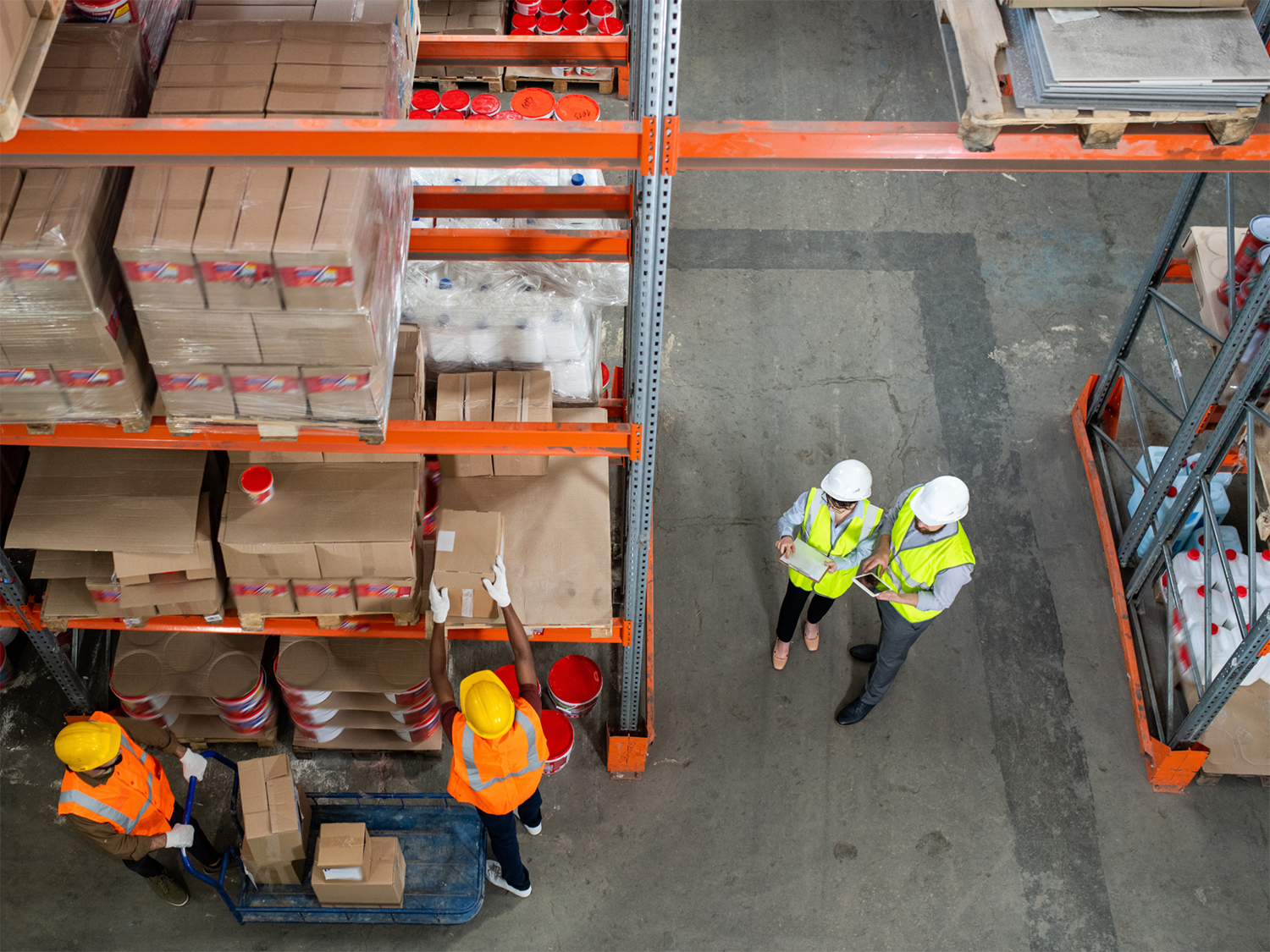 Two workers in yellow safety vests and helmets handling boxes on shelves and a cart in a warehouse, observed from above by two supervisors in white helmets and yellow vests reviewing documents and a tablet on the warehouse floor.