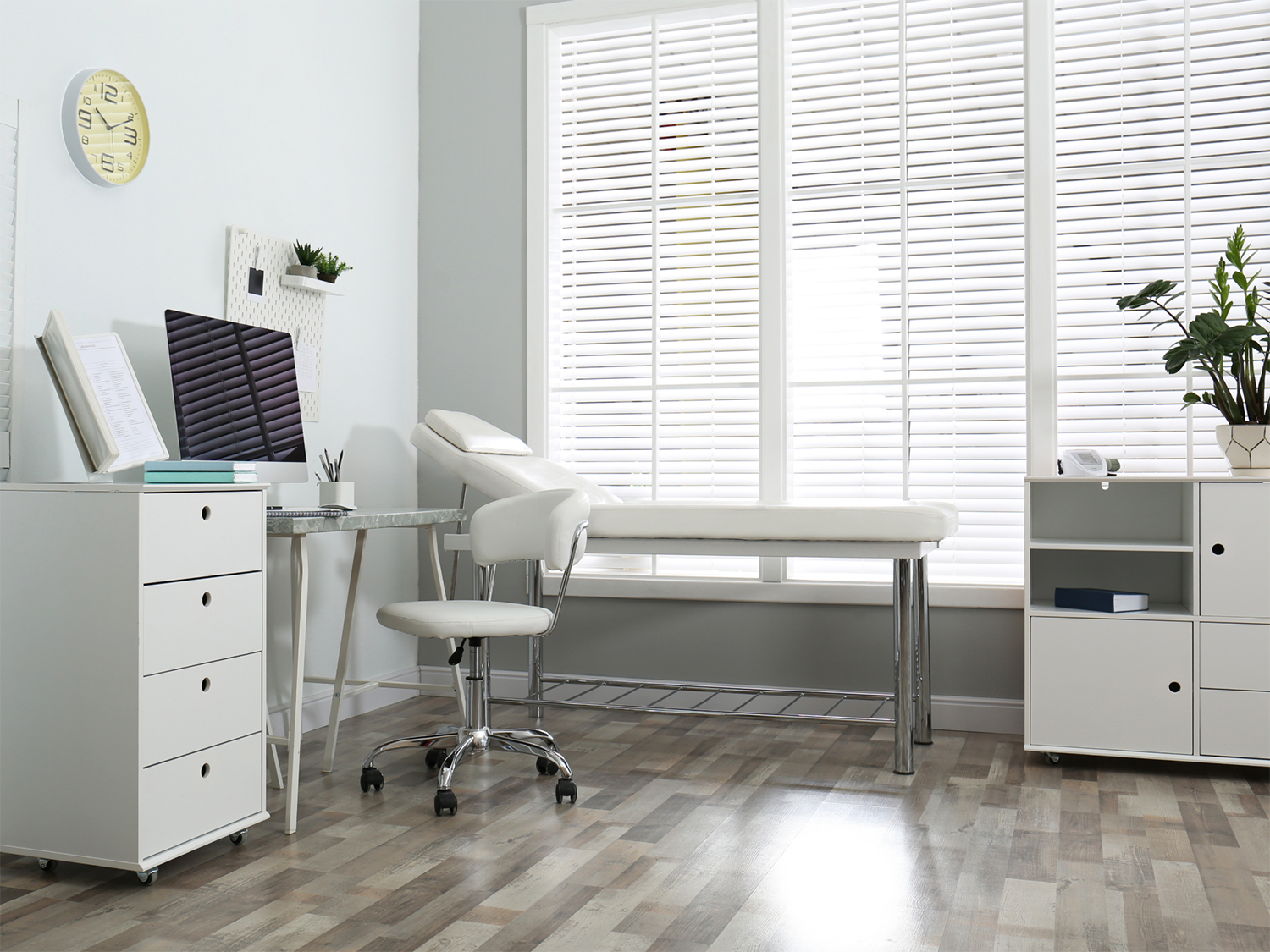 Bright medical consultation room with a white examination table near large windows with white blinds, a rolling white stool, a desk with a computer monitor, and white cabinetry with a plant and a blood pressure monitor on top.