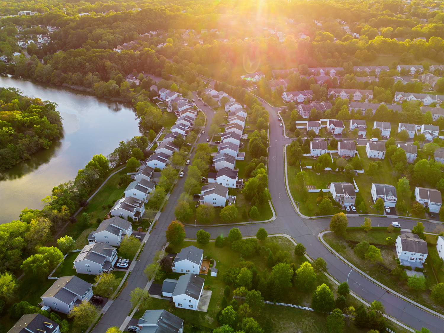 Aerial view of a quiet residential neighborhood with rows of houses, green trees, and a river on the left, bathed in warm sunlight.