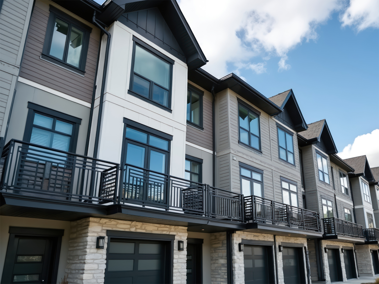 Row of modern townhome community with stone lower walls, large dark framed windows, black metal balconies, and garage doors under a blue sky with scattered clouds.