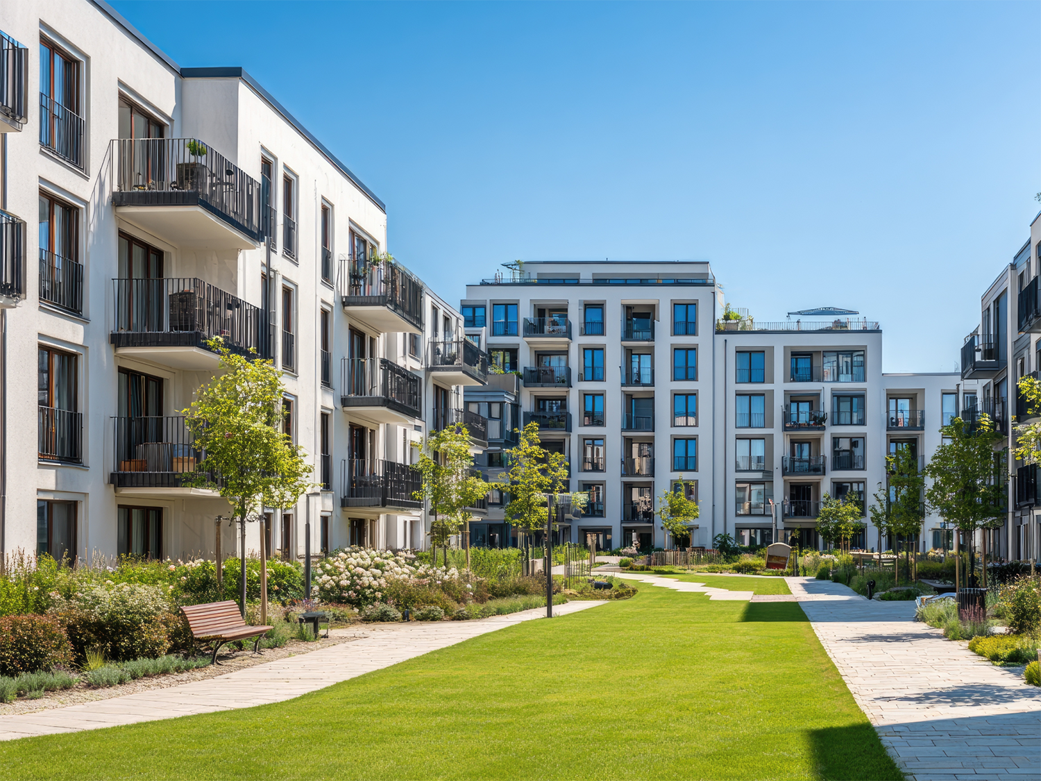 Modern white condominium buildings with multiple balconies surrounding a landscaped courtyard featuring green grass, trees, flowering shrubs, benches, and paved walkways under a clear blue sky.