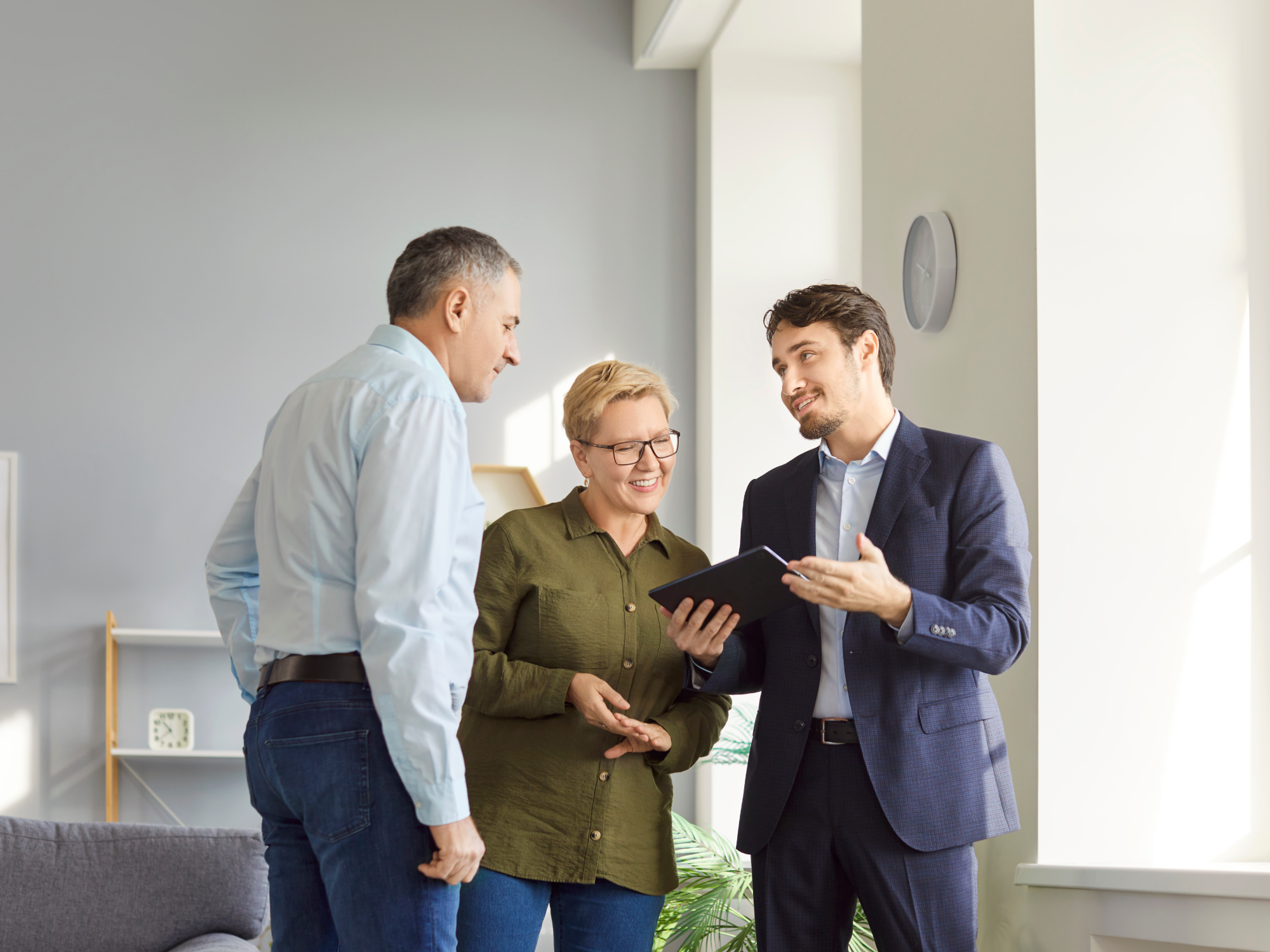 A man in a dark suit shows something on a tablet to an older couple standing beside him, all smiling and engaged in a bright, modern living room.