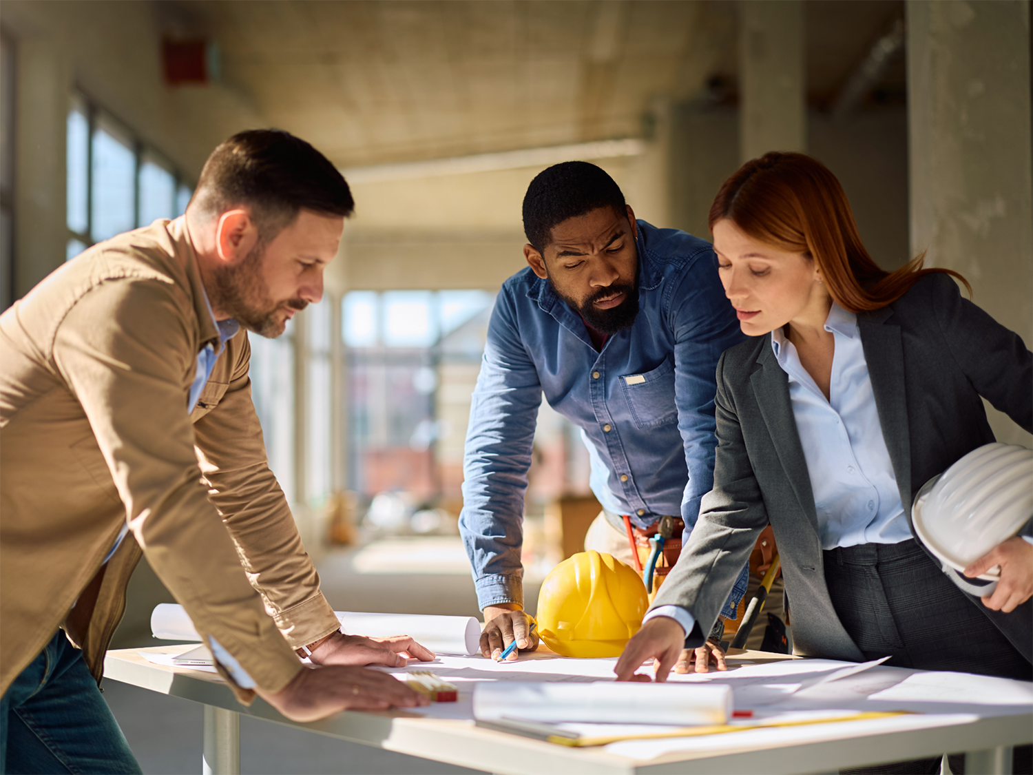 Three professionals, two men and one woman, closely examining blueprints on a table in a spacious, well-lit construction site; the woman holds a white hard hat while a yellow hard hat rests on the table.