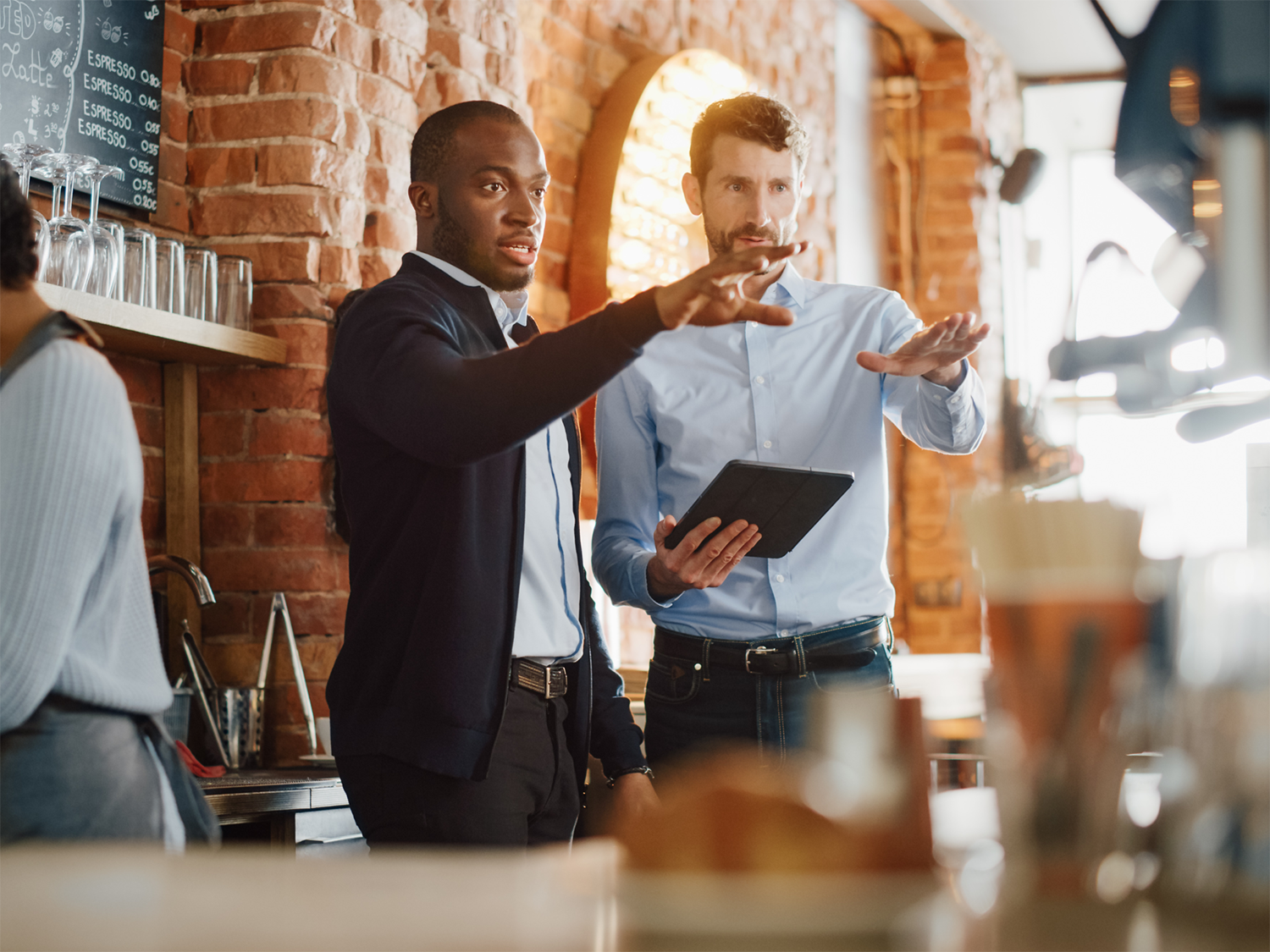 Two men in a café or restaurant are discussing something while one holds a tablet and both gesture with their hands, standing near a brick wall with shelves holding glasses and a chalkboard menu.