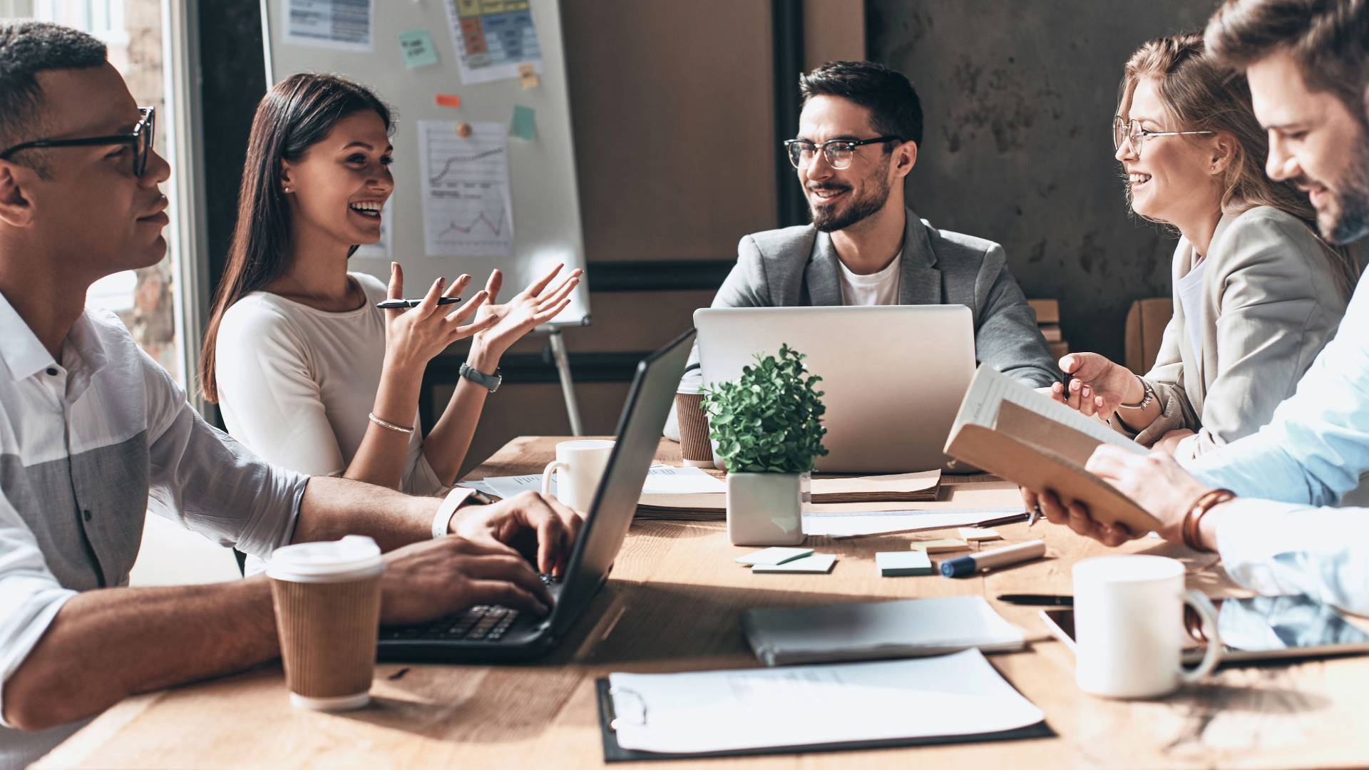 Five smiling professionals sitting around a wooden table in a meeting room, with laptops, coffee cups, documents, and a potted plant on the table, discussing and collaborating, with a whiteboard and window in the background.